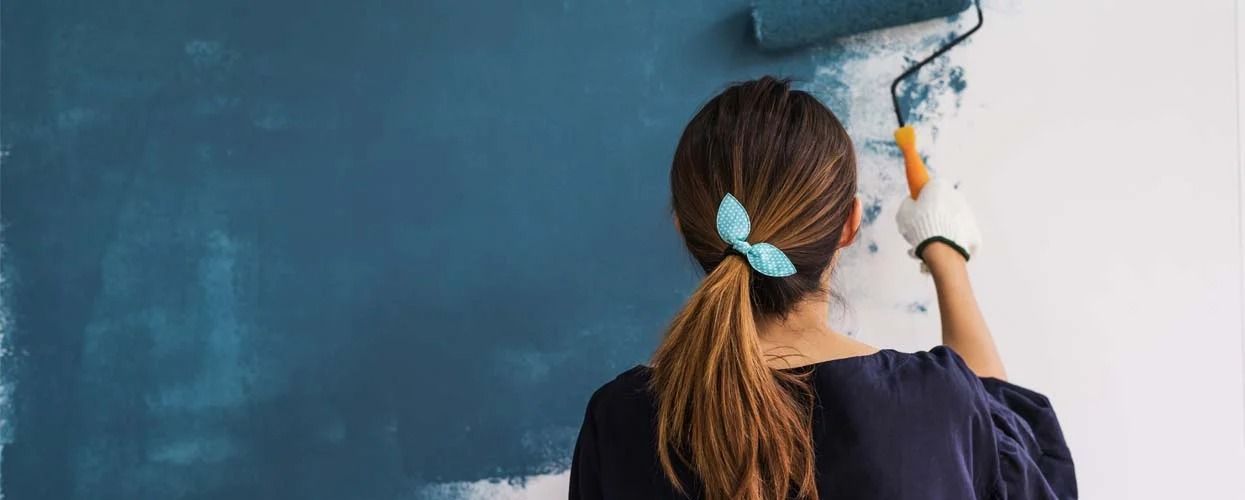 A person with their hair tied back in a ponytail paints a white wall with a roller using deep teal paint.