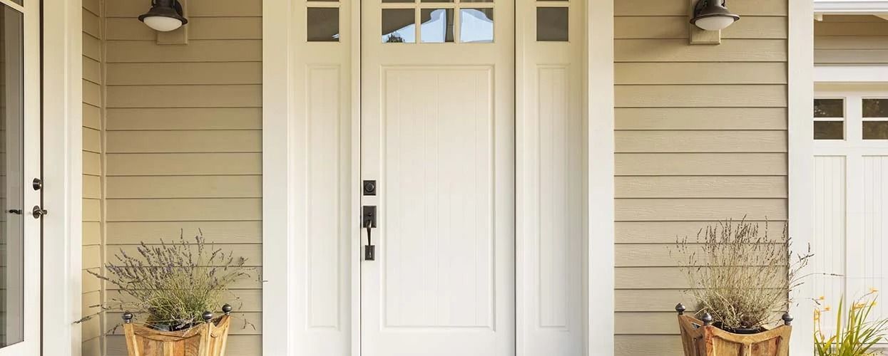 A light-colored front door with sidelights and a transom window, flanked by two potted plants on a beige-sided porch.