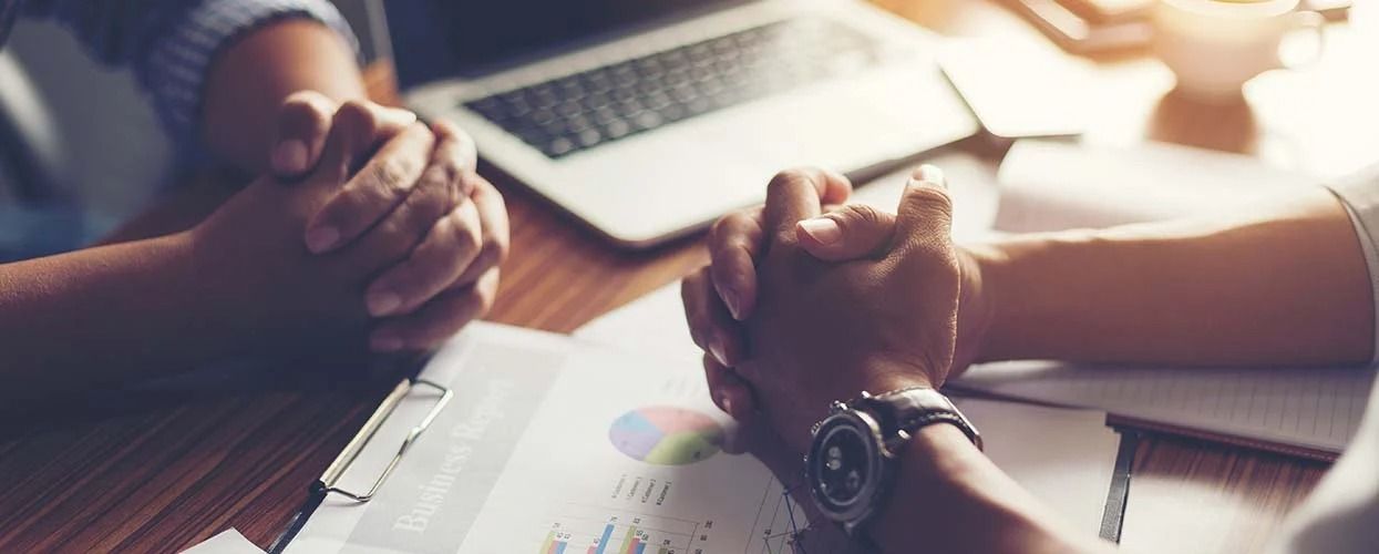 Two people sit at a table with hands clasped, reviewing business documents and a laptop in a brightly lit office.