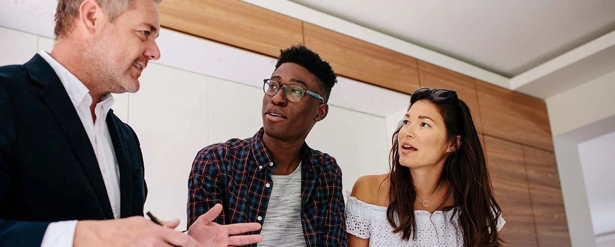 A real estate agent gestures while speaking to two people in a modern kitchen, likely discussing a property.