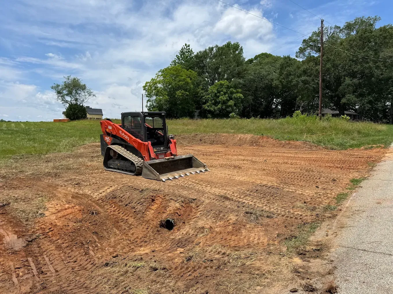 Orange skid steer on a cleared plot of dirt, trees and sky in the background.