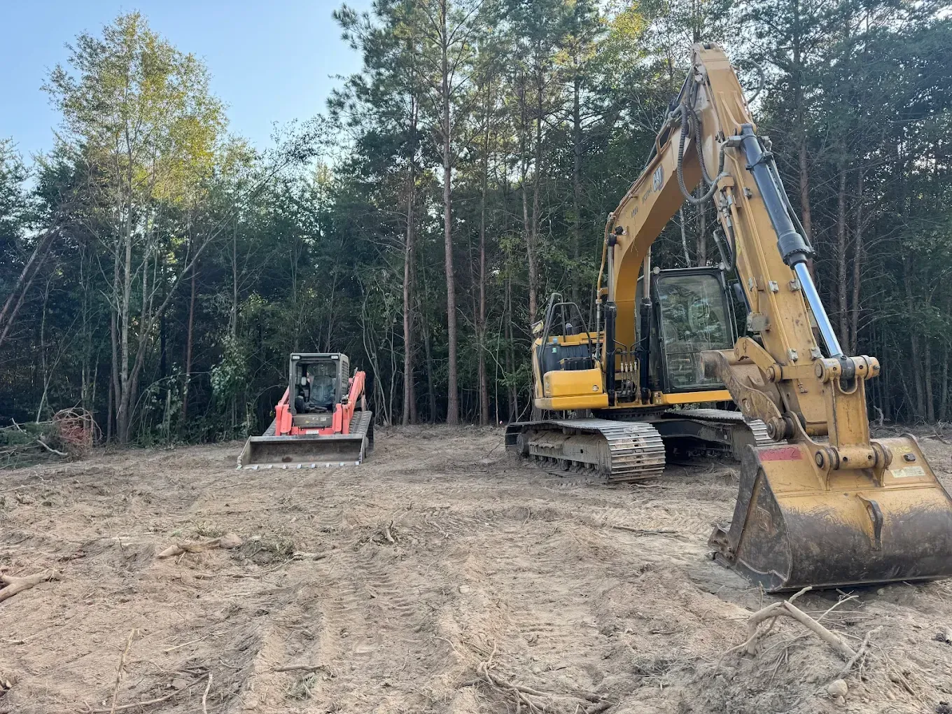 Excavator and skid steer on a cleared plot of land, preparing for construction. Forest background.