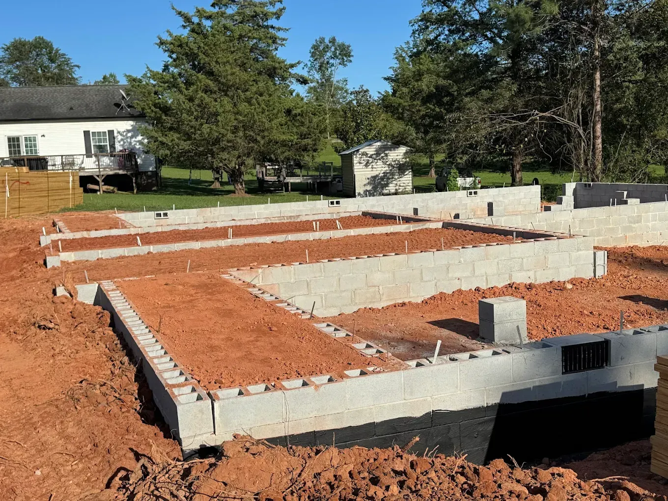 Construction site: Concrete block foundation framework, red dirt, blue sky, and a house in the background.