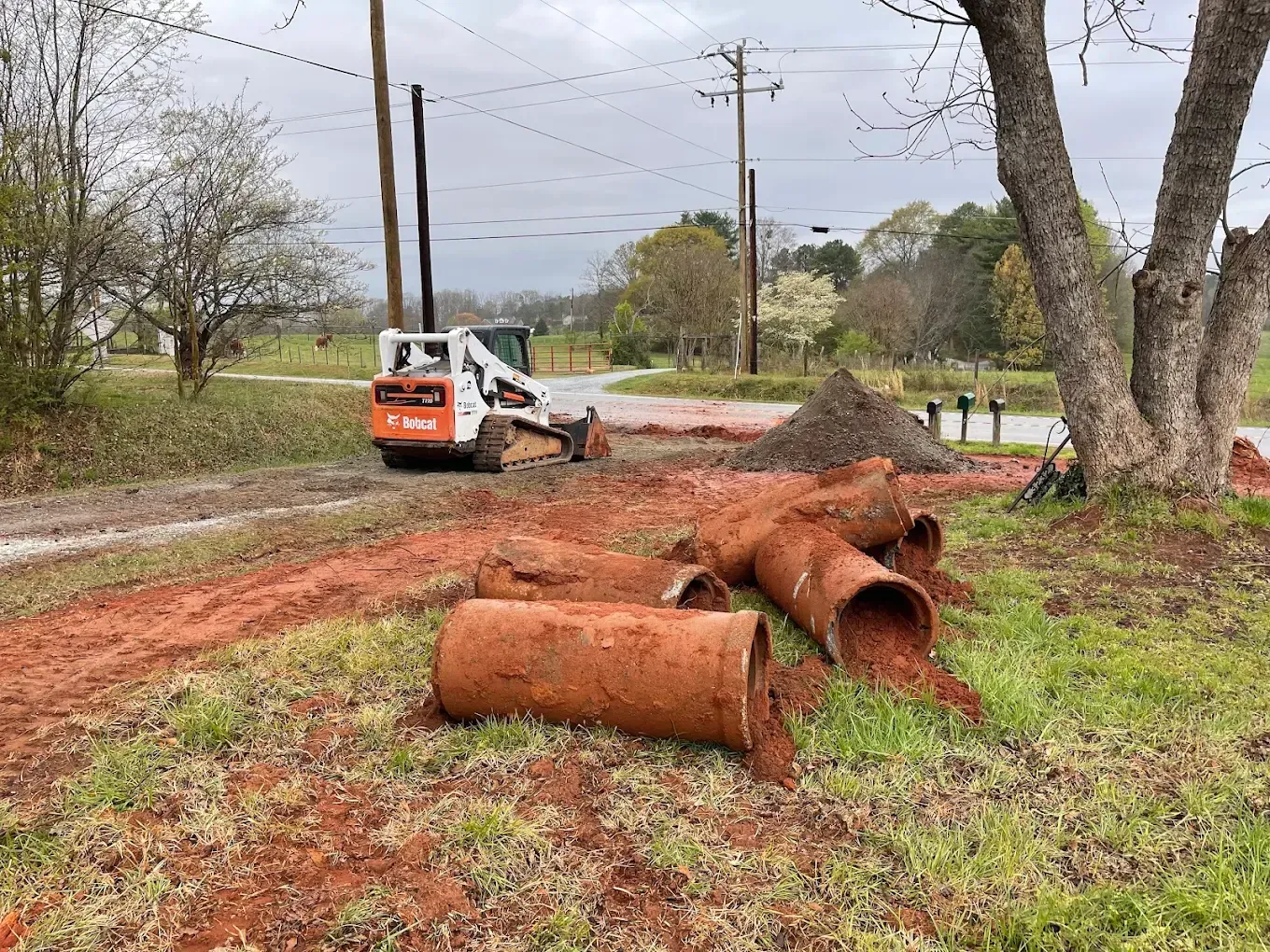 A Bobcat excavator and large pipes on a muddy construction site near a road and power lines.