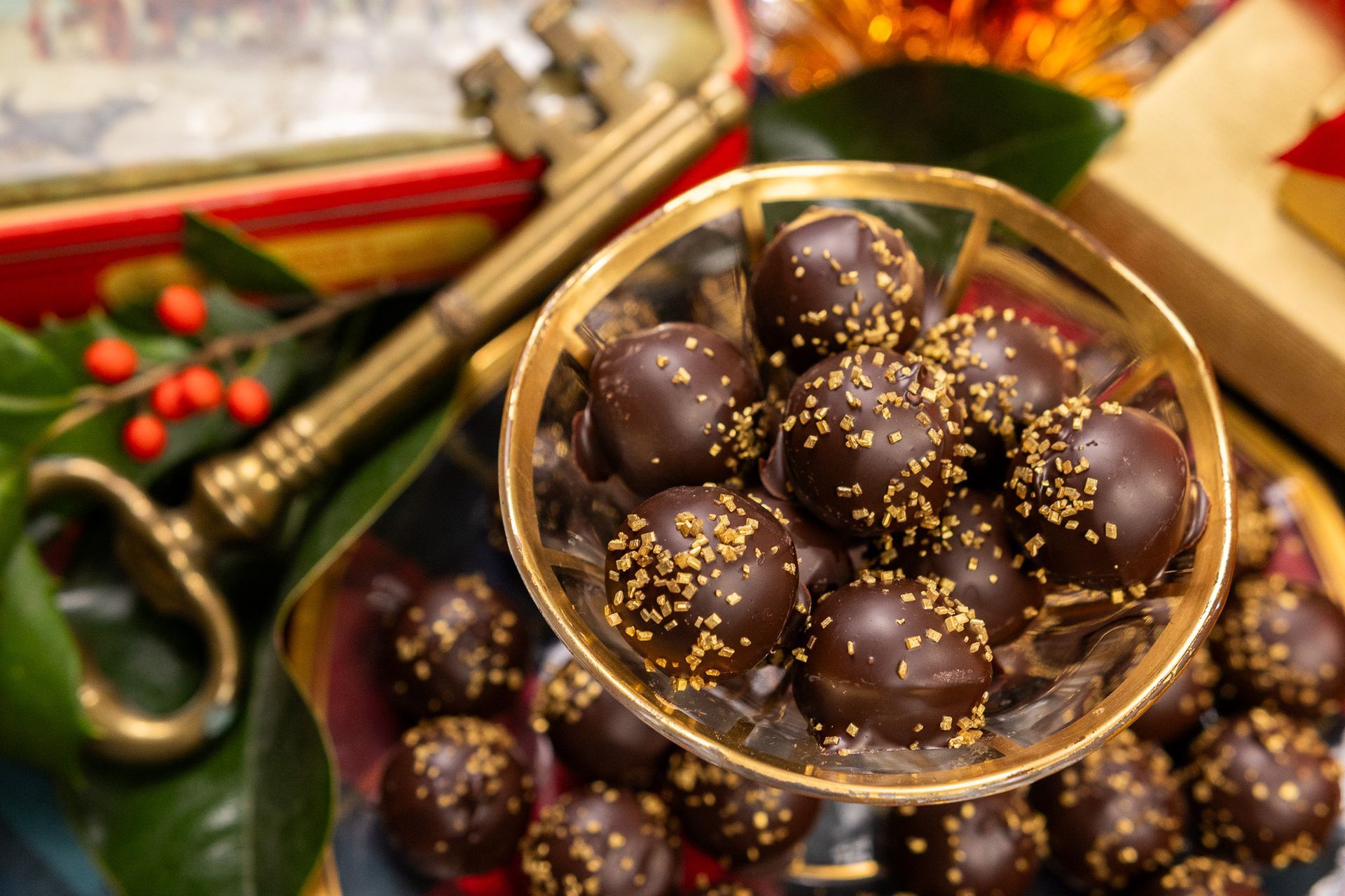 A decorative bowl filled with dark chocolate truffles topped with gold dust, resting near an antique brass key and holly.
