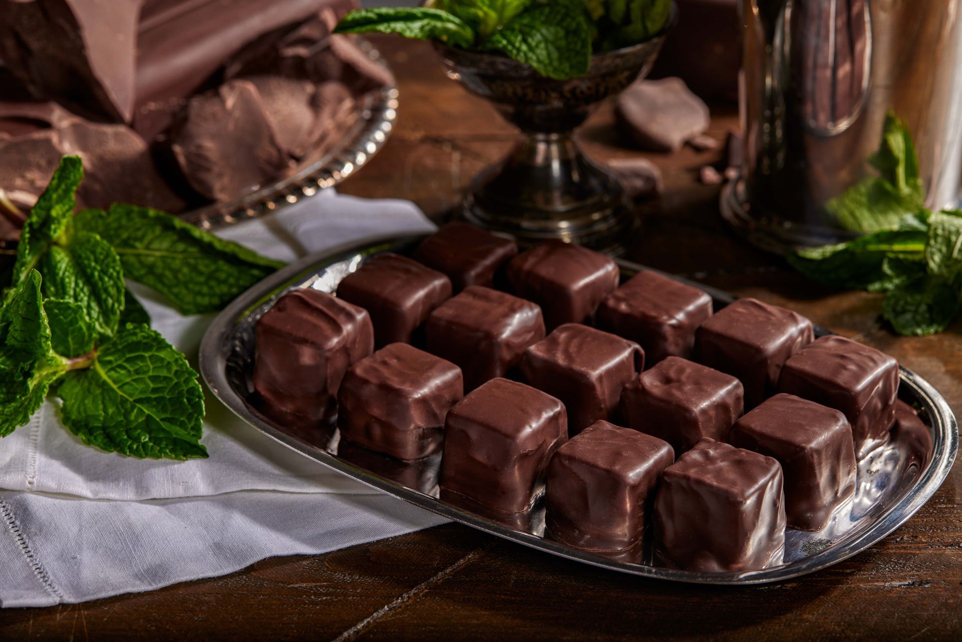 Chocolate mint candies on a silver platter, with mint leaves and chocolate pieces nearby.