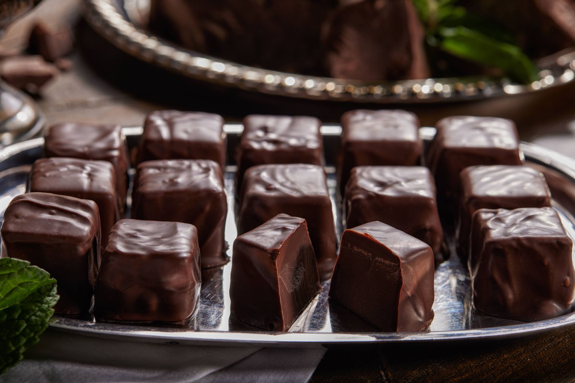 Chocolate mint candies on a silver platter, with mint leaves and chocolate pieces nearby.