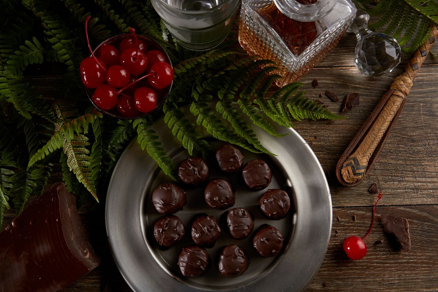 Dark chocolate cherries on a plate with maraschino cherries and whiskey on a wood table with green fern.