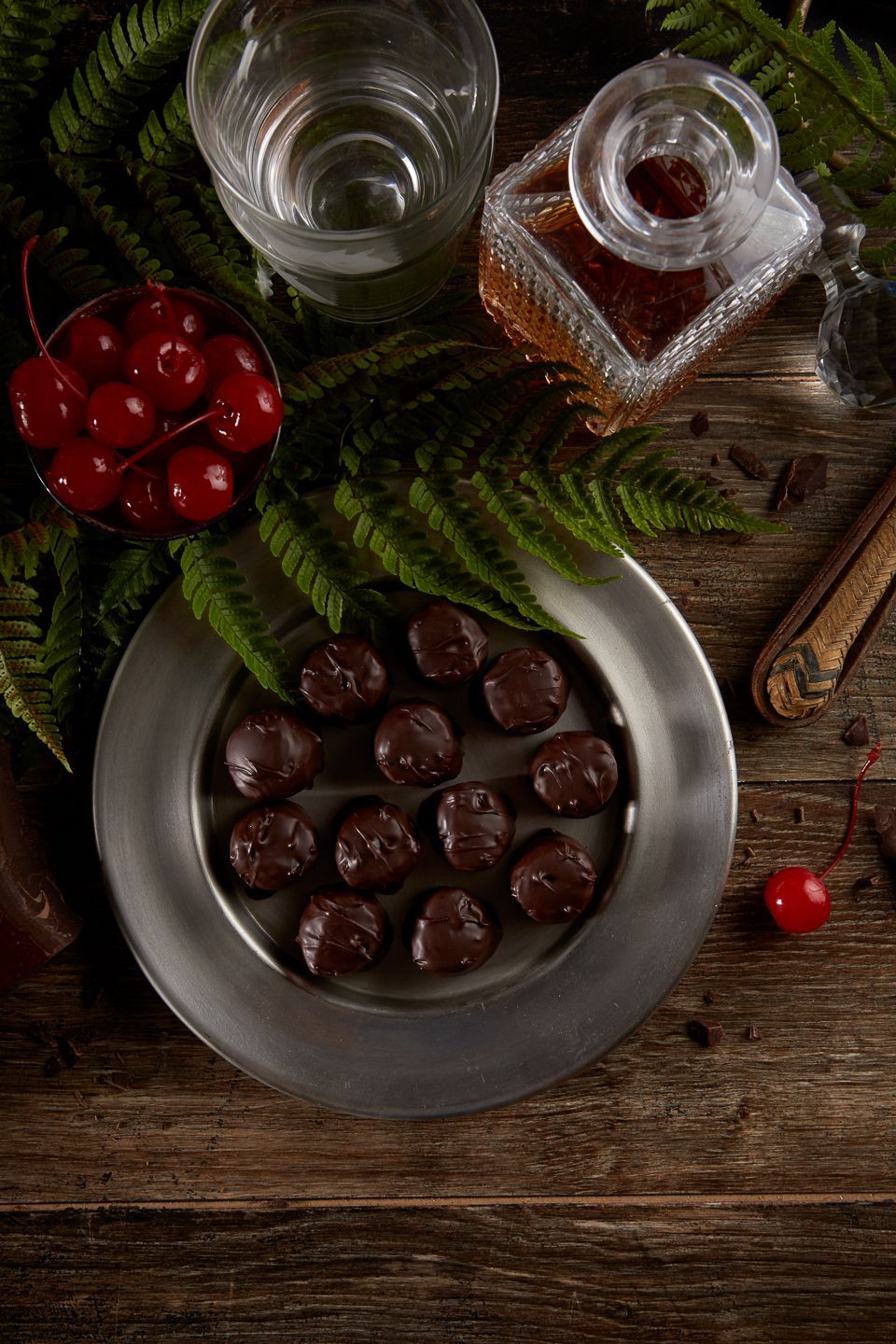 Chocolate-covered cherries on a pewter plate, with fresh cherries, a liquor bottle, glass, and wood table.