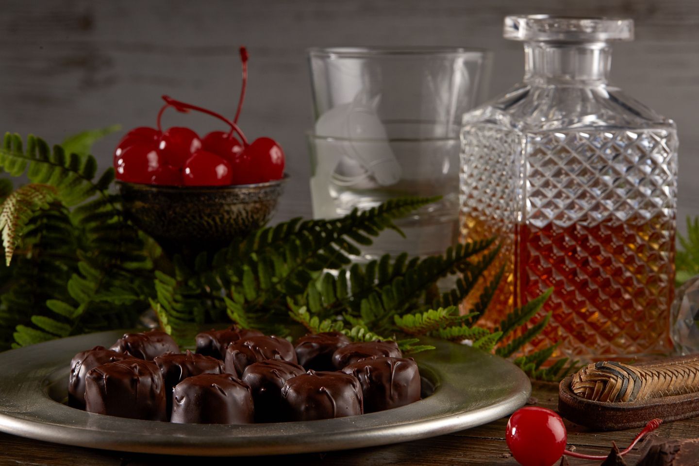 Plate of chocolate candies, bowl of cherries, and decanter with drink set among greenery.