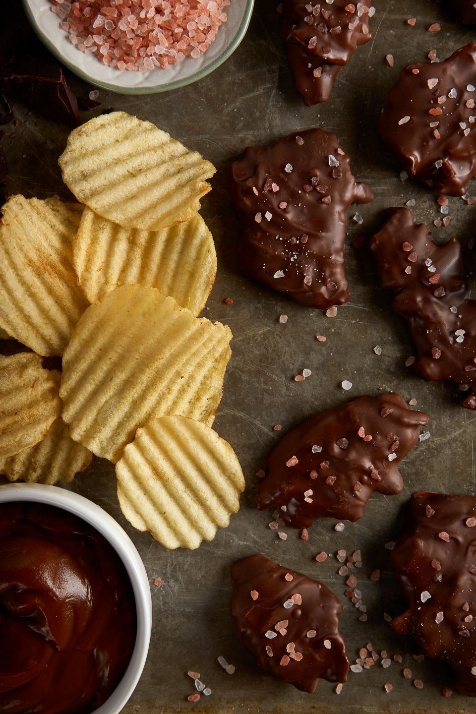 Chocolate-covered snacks sprinkled with salt next to wavy potato chips and bowls of chocolate sauce and pink salt.
