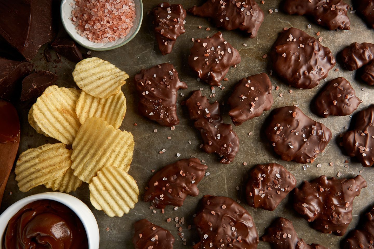Chocolate-covered potato chips in a white bowl with a silver rim. Dark chocolate bar and potato chips visible.