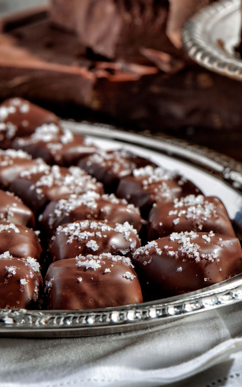 A silver platter holding rows of chocolate-covered sea salt caramels, with dark chocolate chunks in the background.