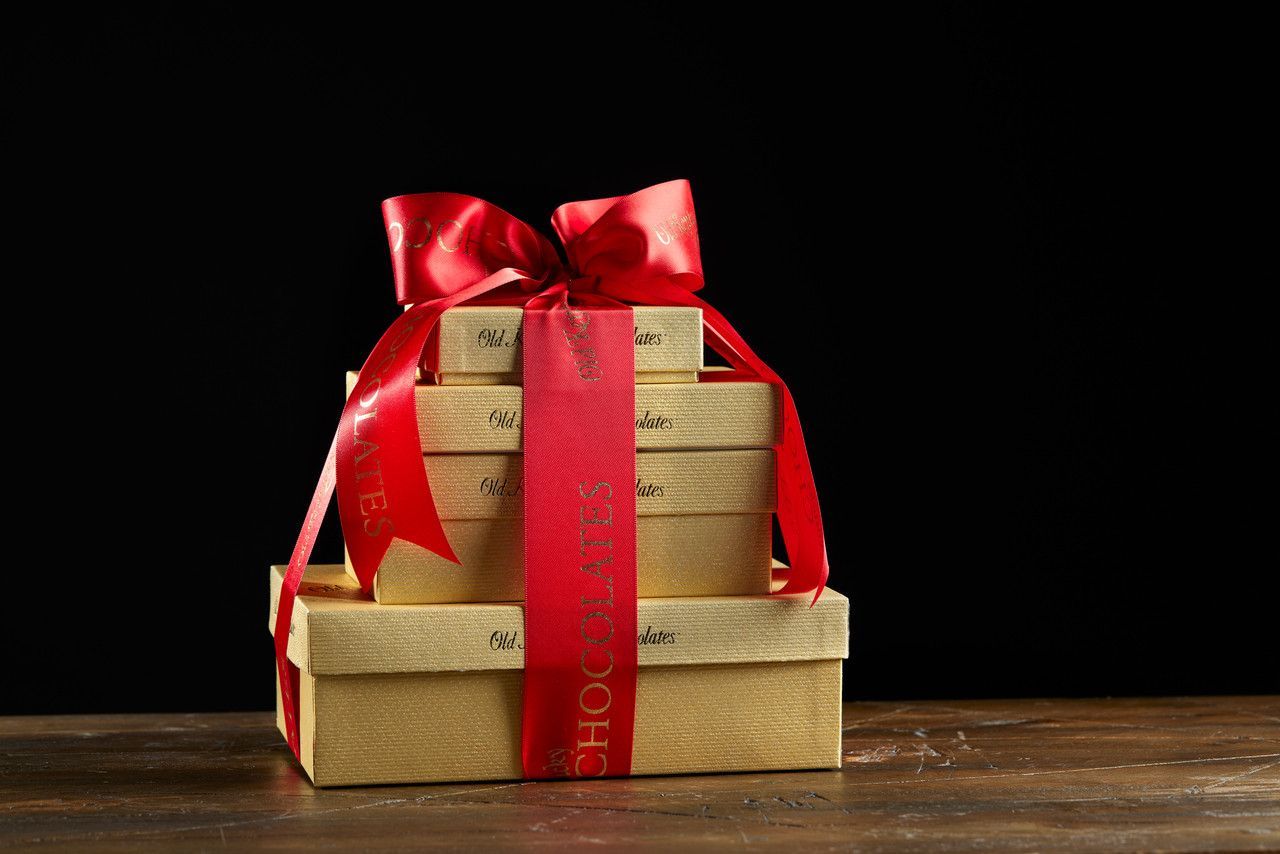 Gold gift boxes stacked, tied with a red ribbon and bow. The boxes sit on a wooden surface against a black backdrop.