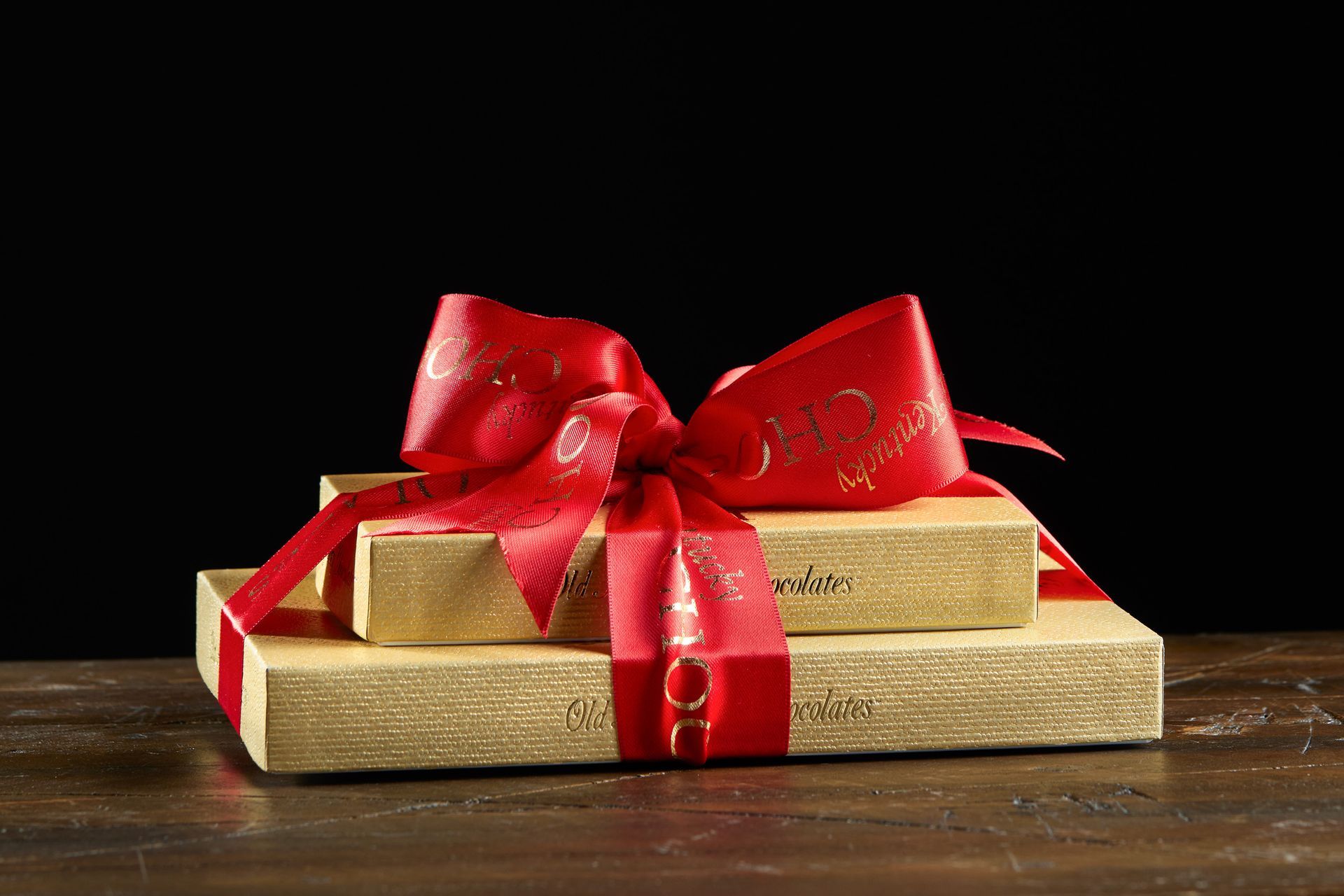 Gold wrapped gifts stacked with a red ribbon and bow, on a wooden table. Black background.