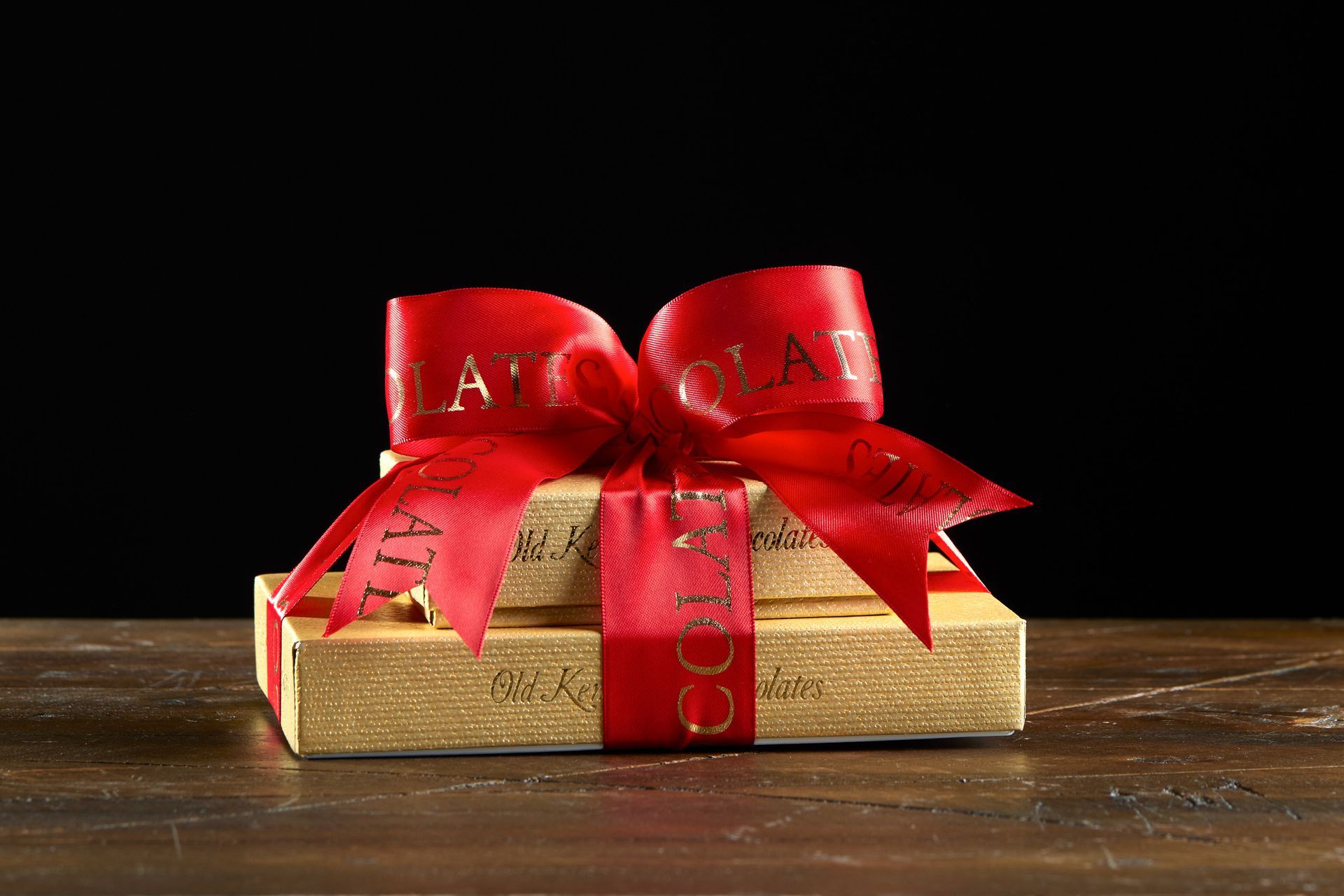 Gift box with gold wrapping, tied with a red ribbon and bow on a wooden surface against a black background.