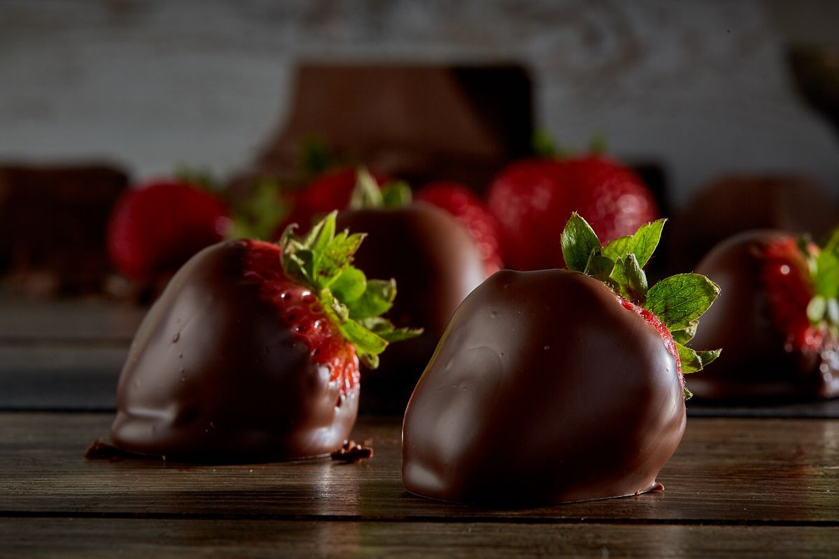 Chocolate-covered strawberries on a wooden surface, with red berries in the background.