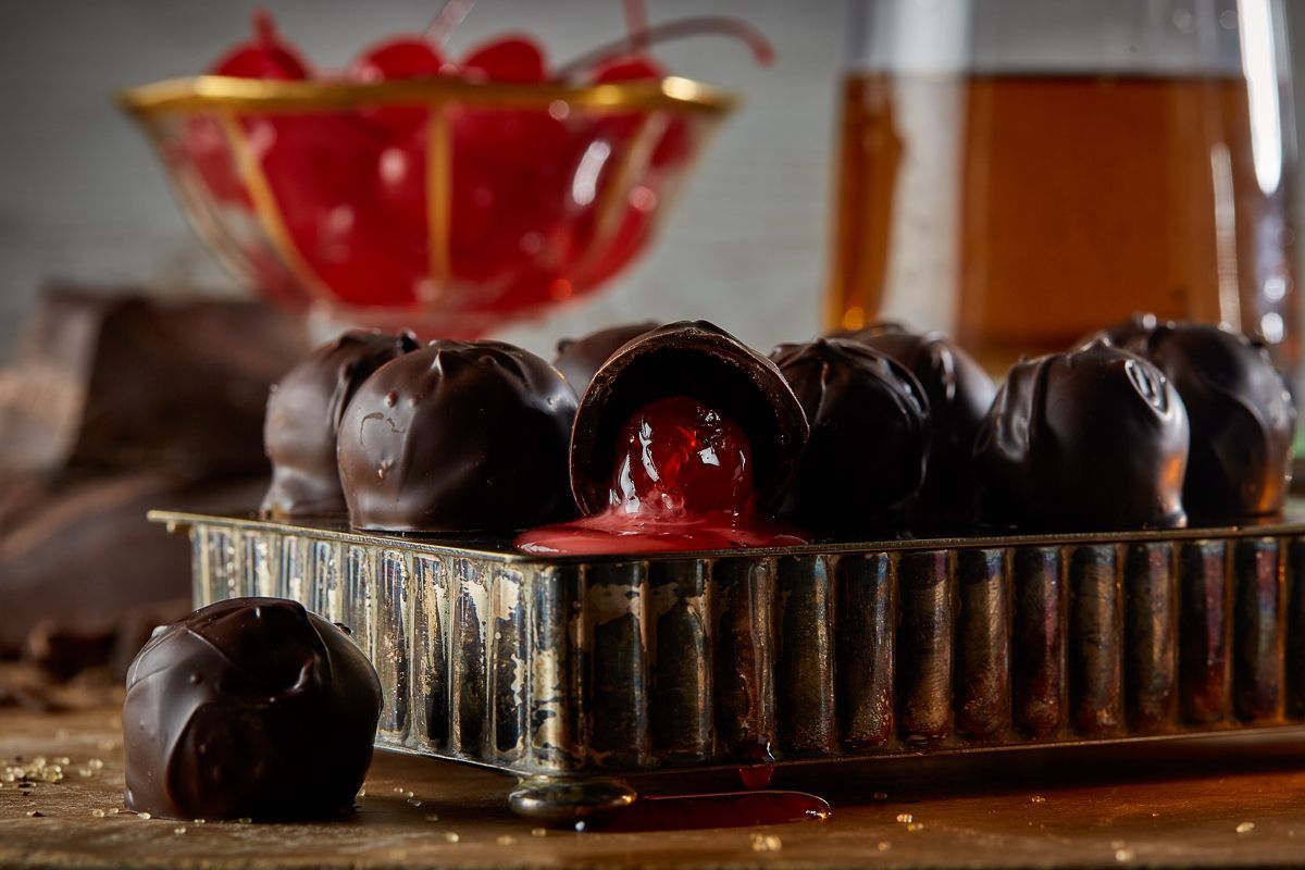 Dark chocolate cherries on a plate with maraschino cherries and whiskey on a wood table with green fern.
