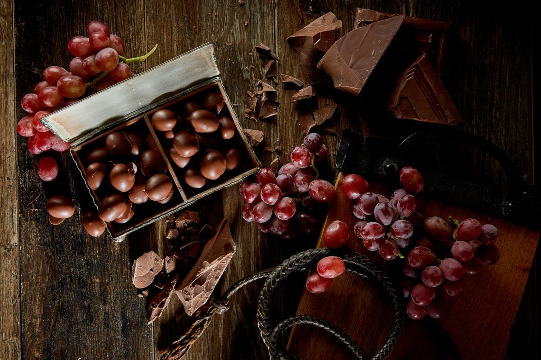 Dark chocolate-covered grapes in a box, scattered on wood table with chocolate chunks and red grapes.