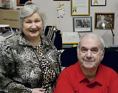 Woman in animal print jacket stands beside a man in a red shirt. They smile in an office setting.