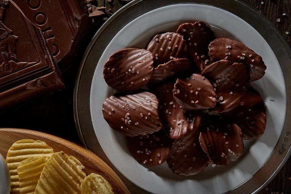 Chocolate-covered potato chips in a white bowl with a silver rim. Dark chocolate bar and potato chips visible.