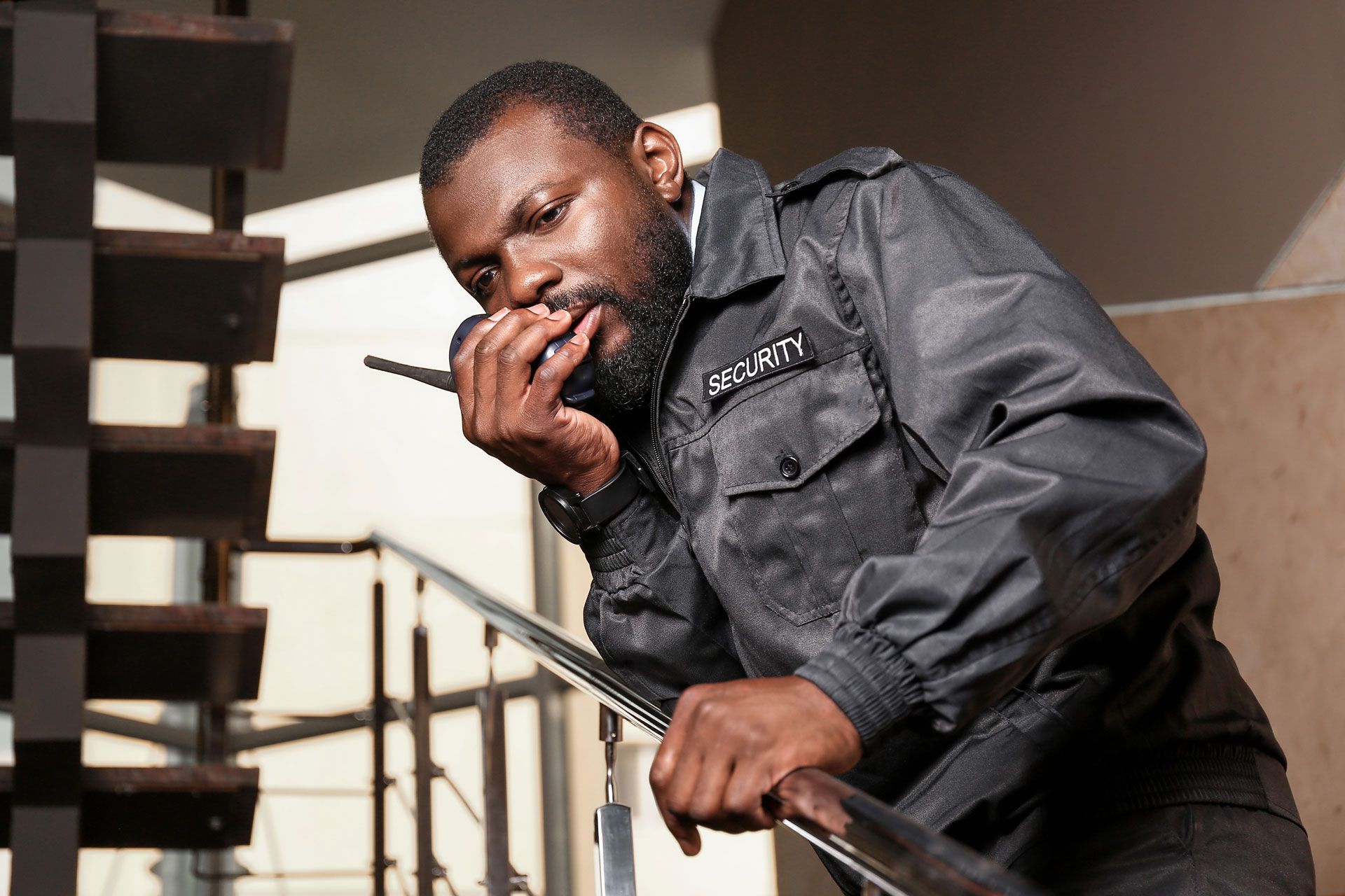 Man in a gray jacket speaking into a walkie-talkie on an indoor stairway.