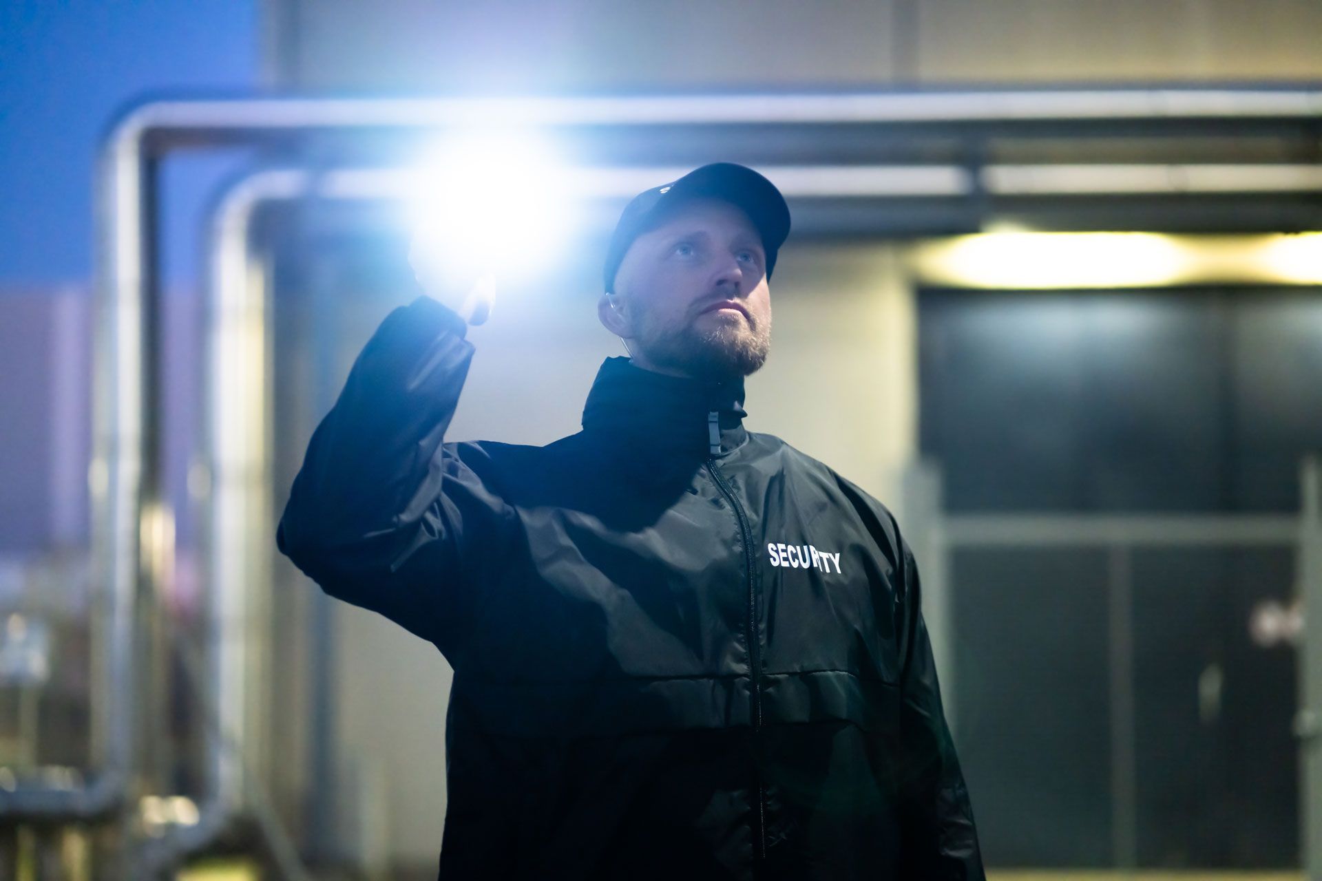 Worker in dark jacket and cap holding up a bright flashlight indoors at night.