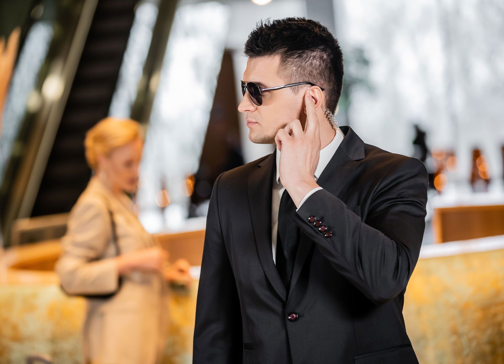 Man in black suit and sunglasses using a smartphone outdoors, with a blurred woman in the background