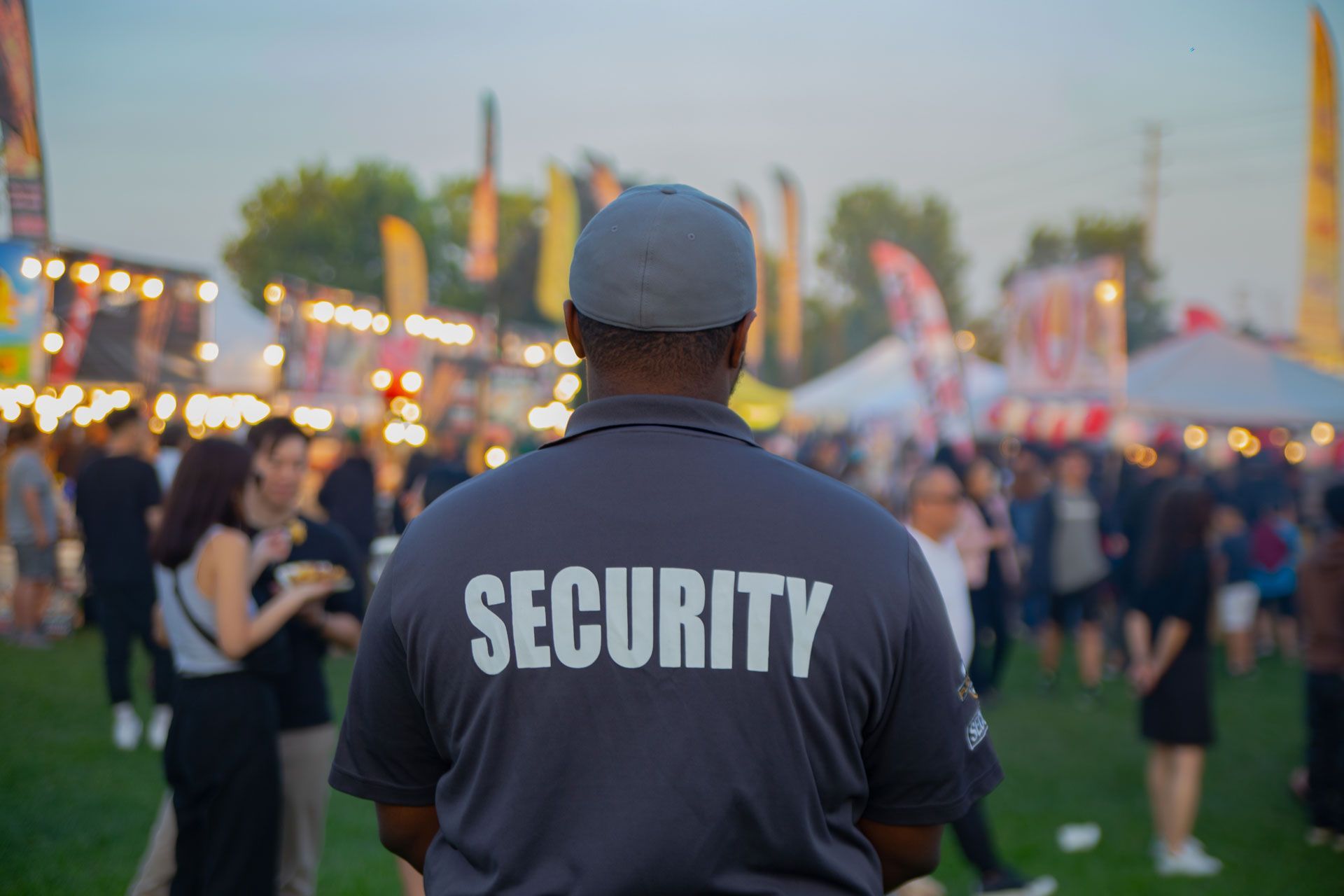 Security guard with “SECURITY” on back, standing at an outdoor crowd event at dusk.