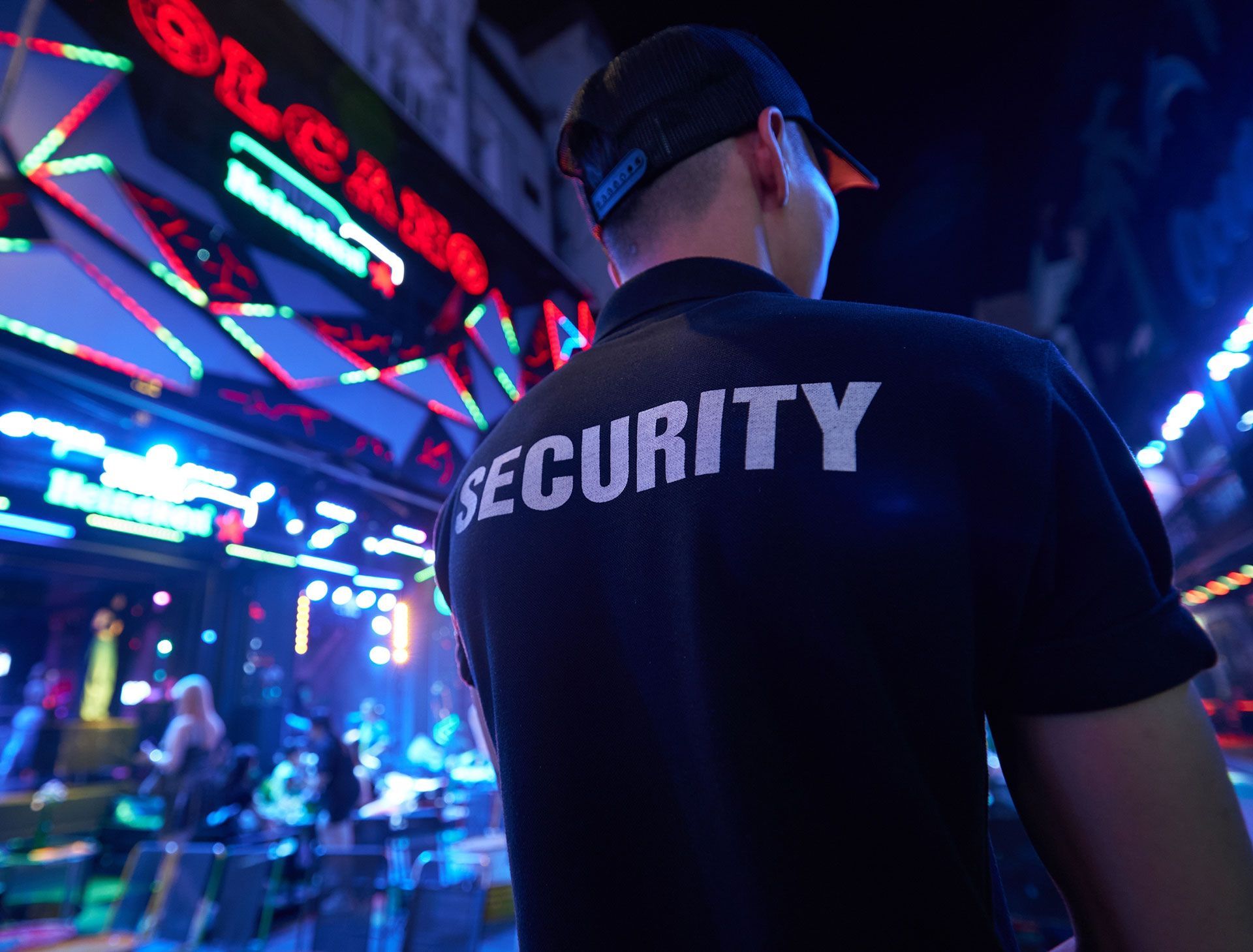 Security guard in black shirt standing in a neon-lit venue, seen from behind.