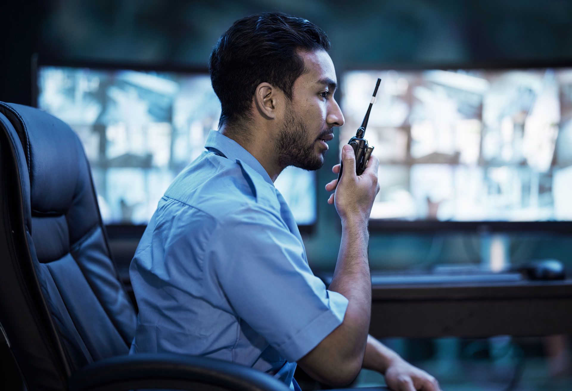 Man in blue shirt speaking into a handheld radio in a control room, seated by multiple monitors.