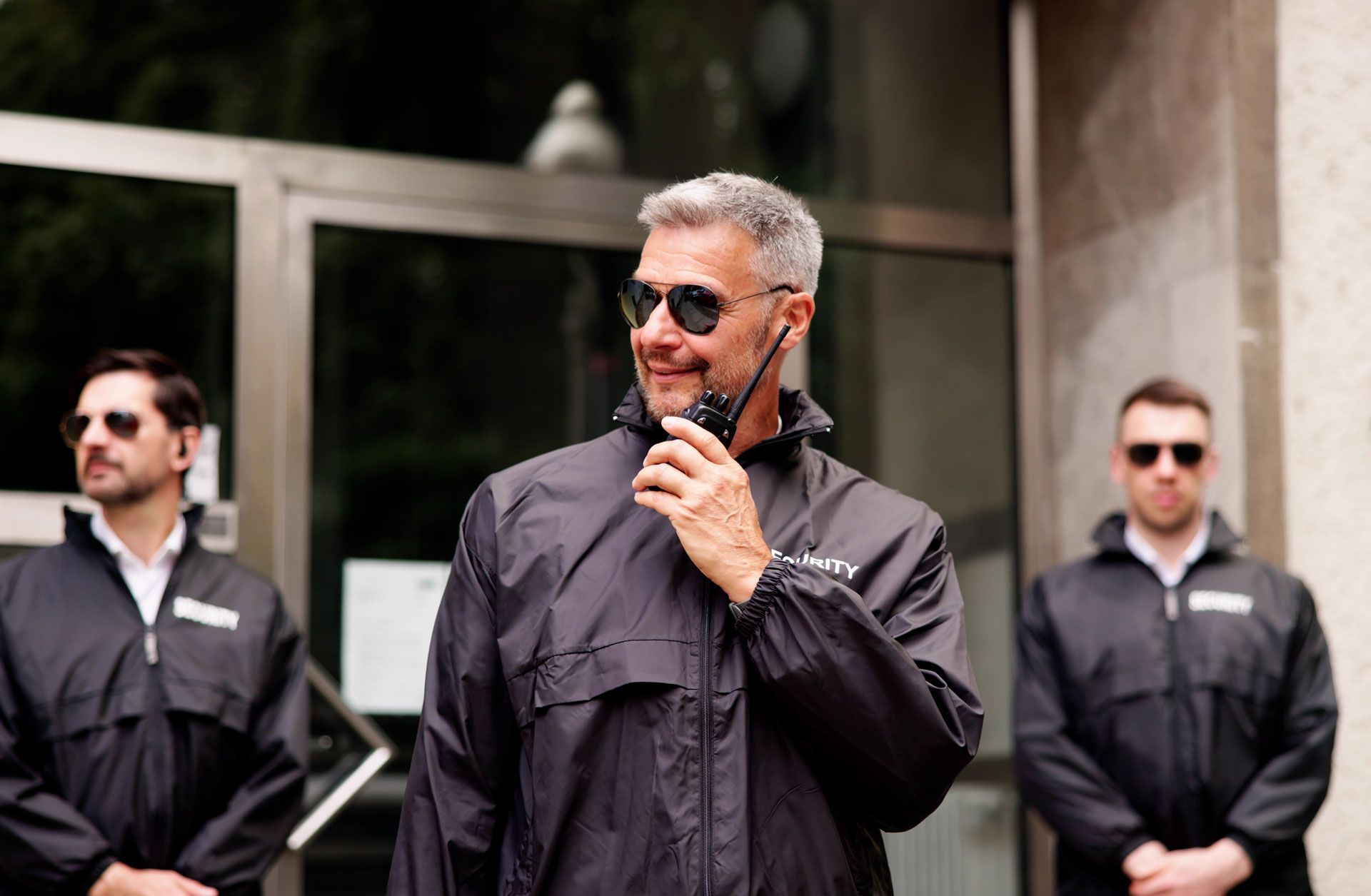 Three men in dark jackets stand outside a building; the center man speaks into a handheld radio.