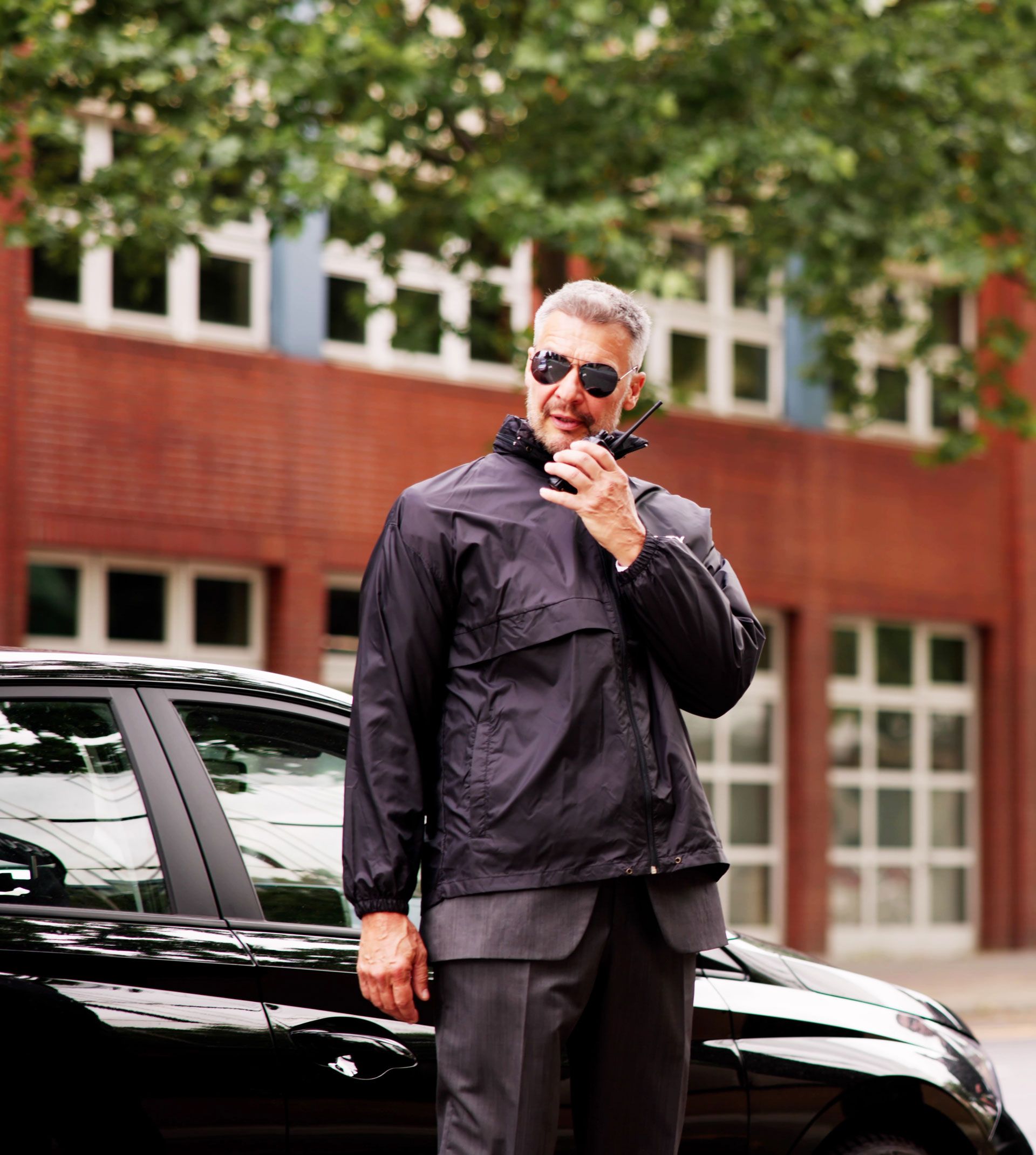 Man in sunglasses speaking into a handheld microphone beside a black car, outdoors in front of a brick building.
