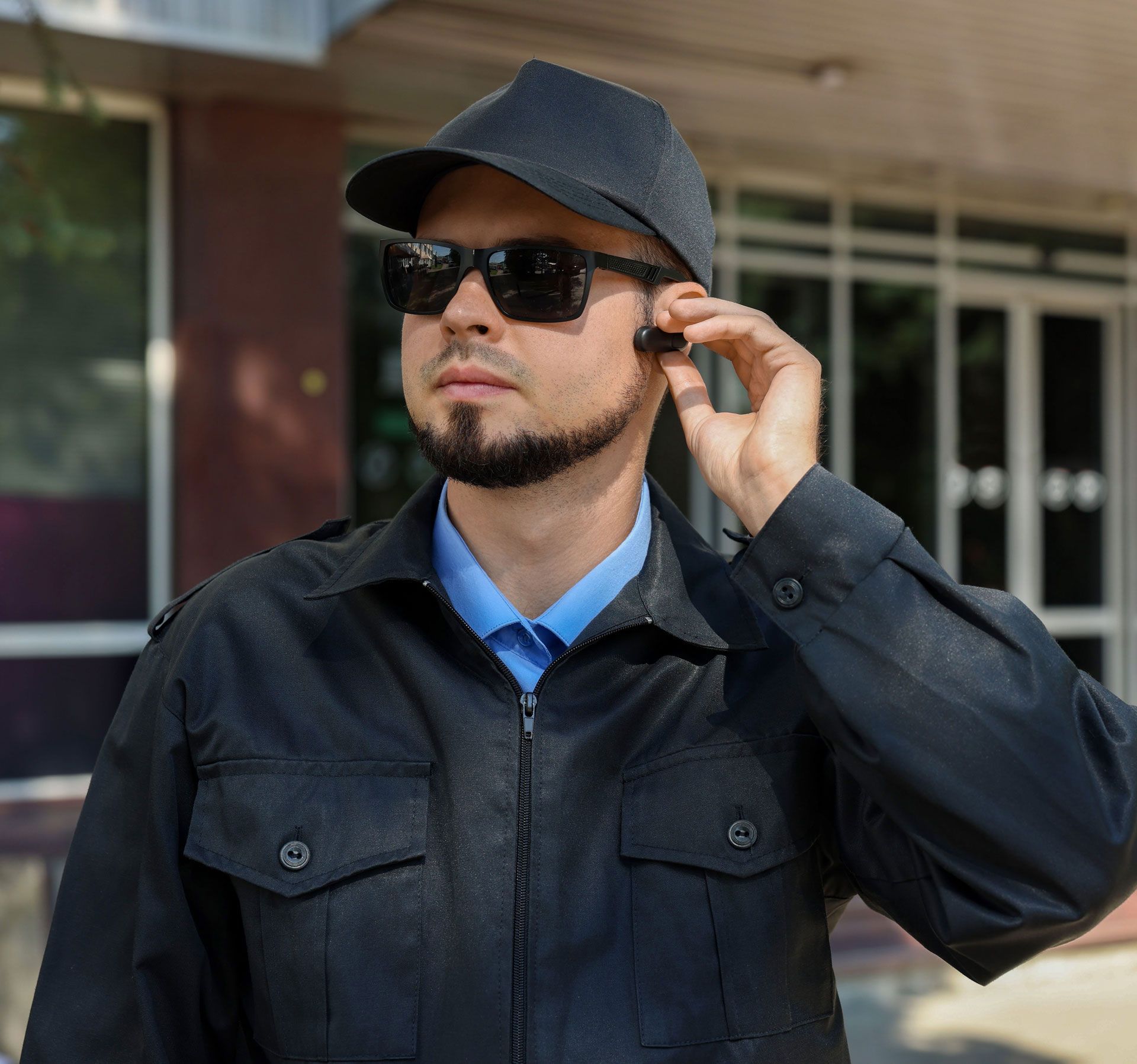 Man in black jacket and cap adjusting sunglasses outside a building