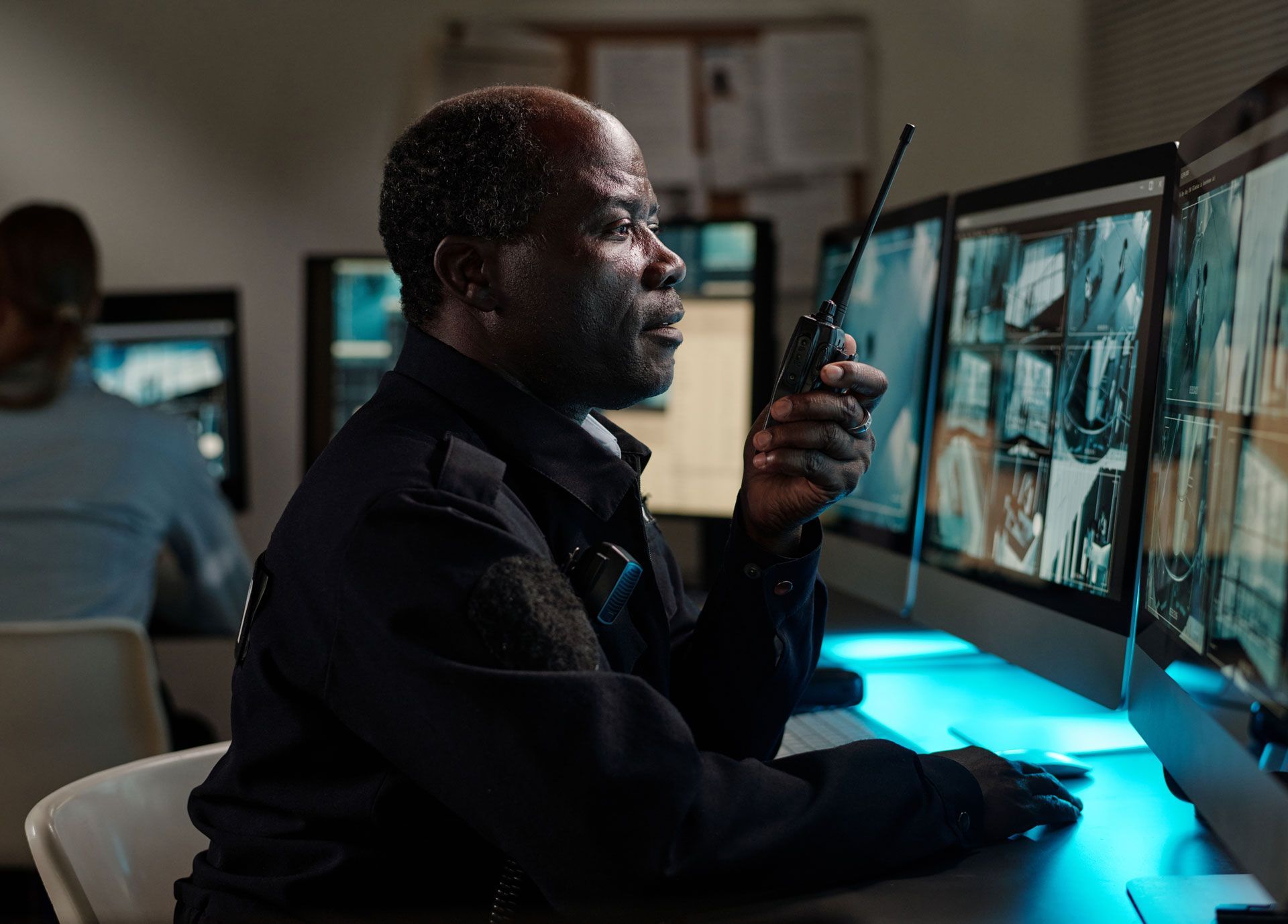 Person using a radio at a desk with dual monitors in a dim control room, lit by blue screen glow