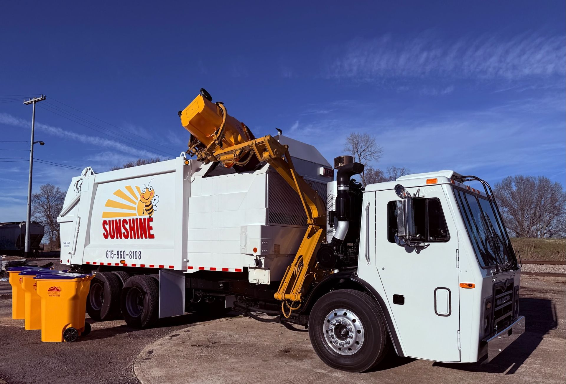 Yellow garbage truck lifting a dumpster into a white refuse truck under a blue sky