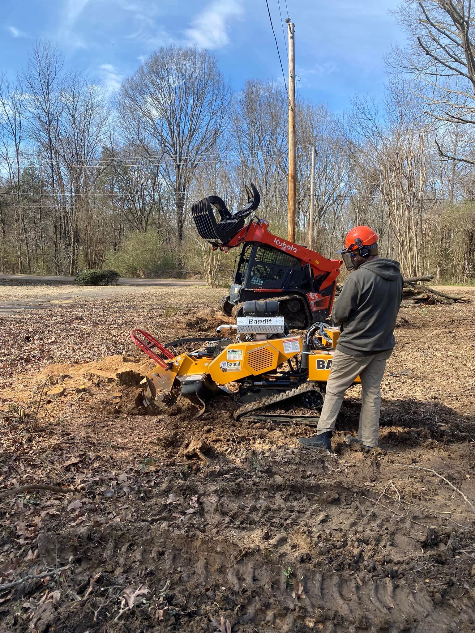 A person in safety gear watches an orange and black forestry machine mulching wood in a wooded area on a sunny day.