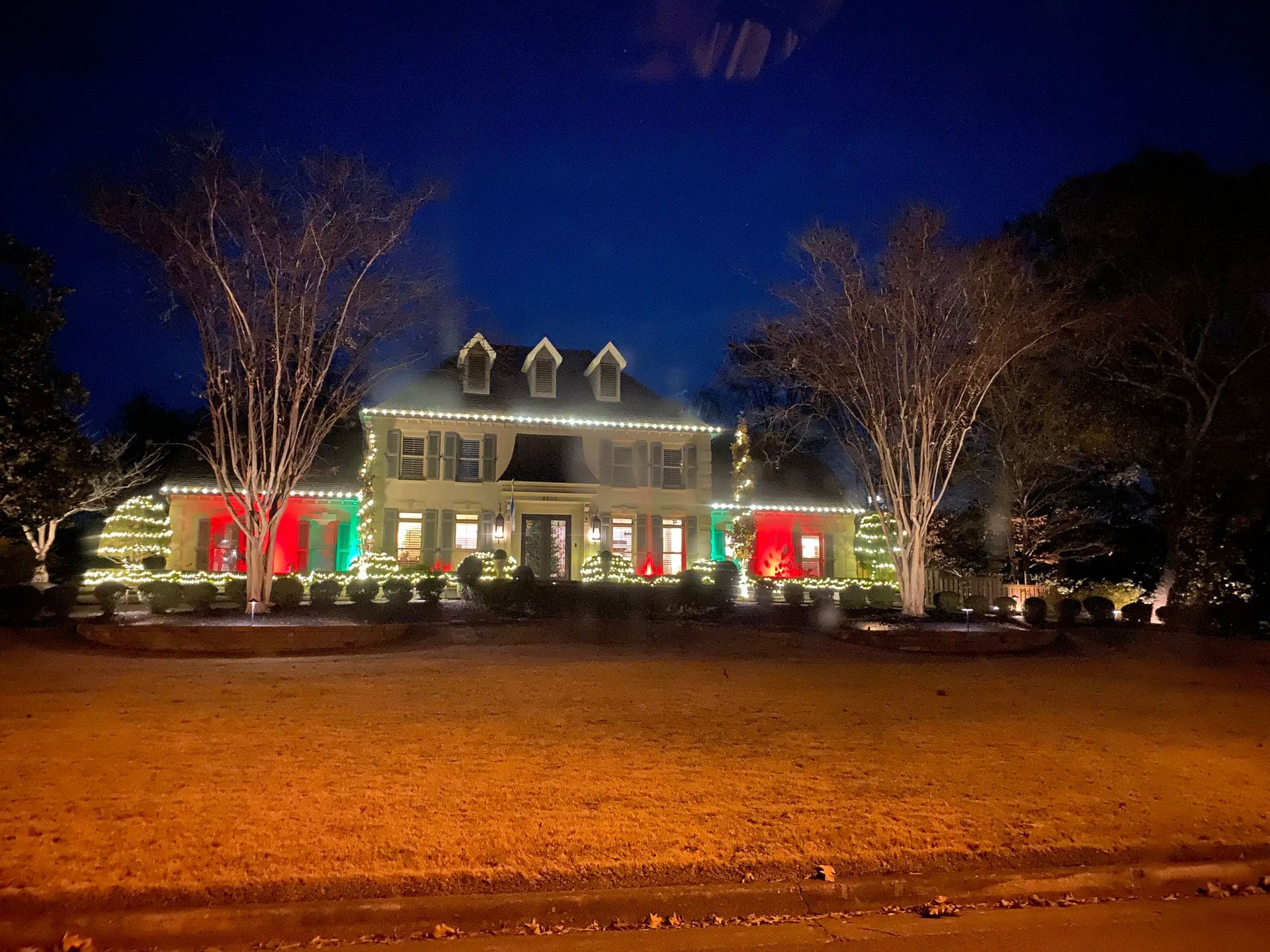 A large house decorated with Christmas lights at night. Red and green accents adorn the facade and trees, and white lights outline the roof.