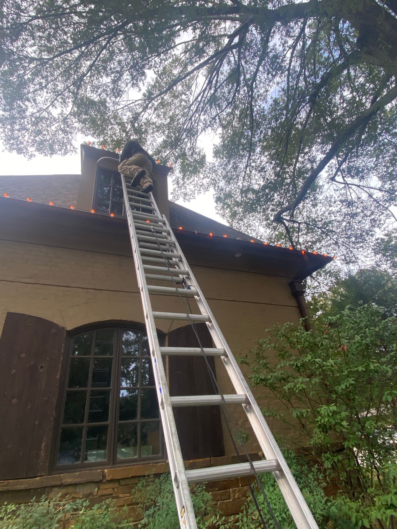 Person on a ladder working on a house's roof near a window. The house has brown shutters and a red tile roof.