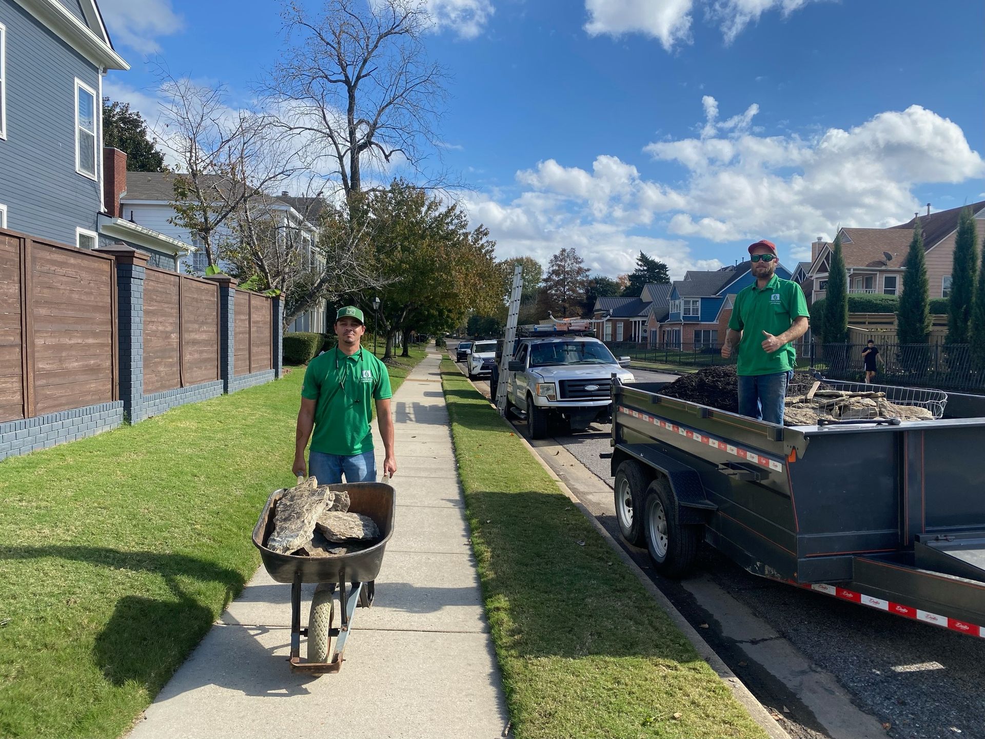 Two men in green shirts and caps remove debris from a sidewalk, using a wheelbarrow and trailer on a sunny residential street.