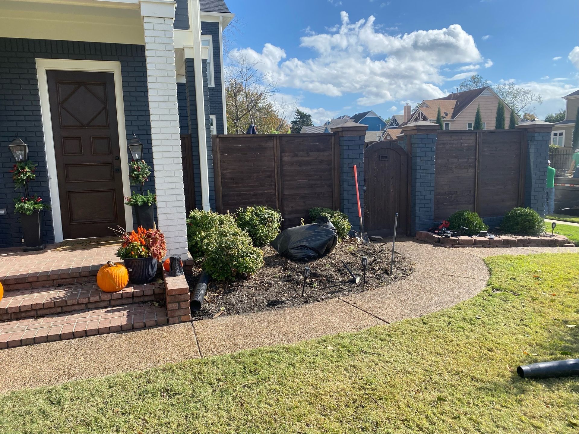 A front yard with a walkway leading to a navy blue house. A wooden fence encloses the garden bed with bushes and a large rock.