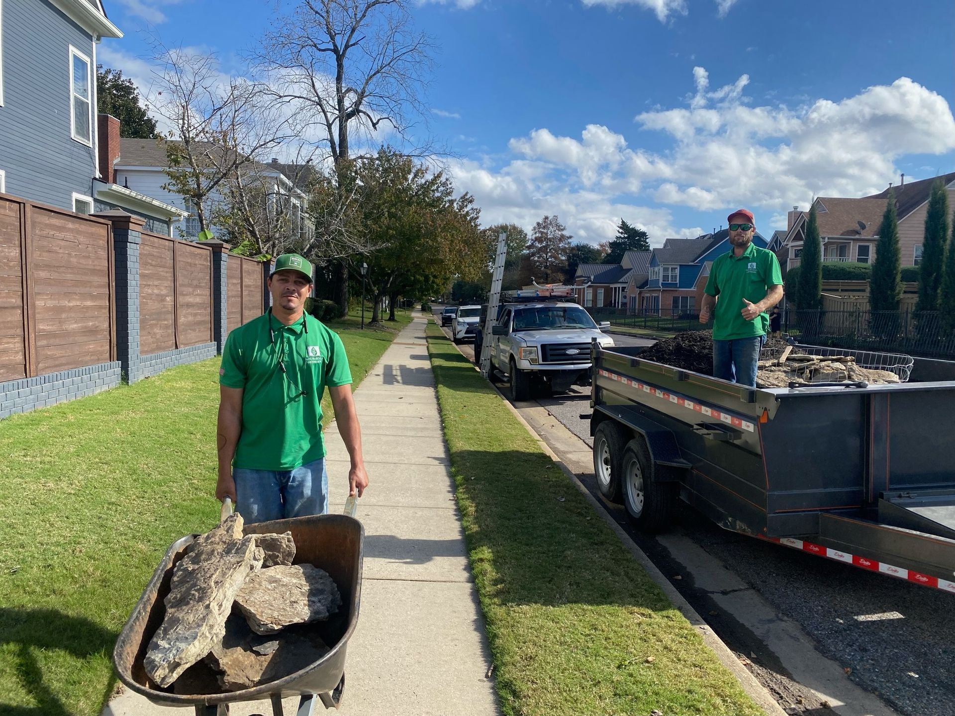 Two men in green shirts working on a sidewalk, one pushing a wheelbarrow, another by a trailer loading debris.