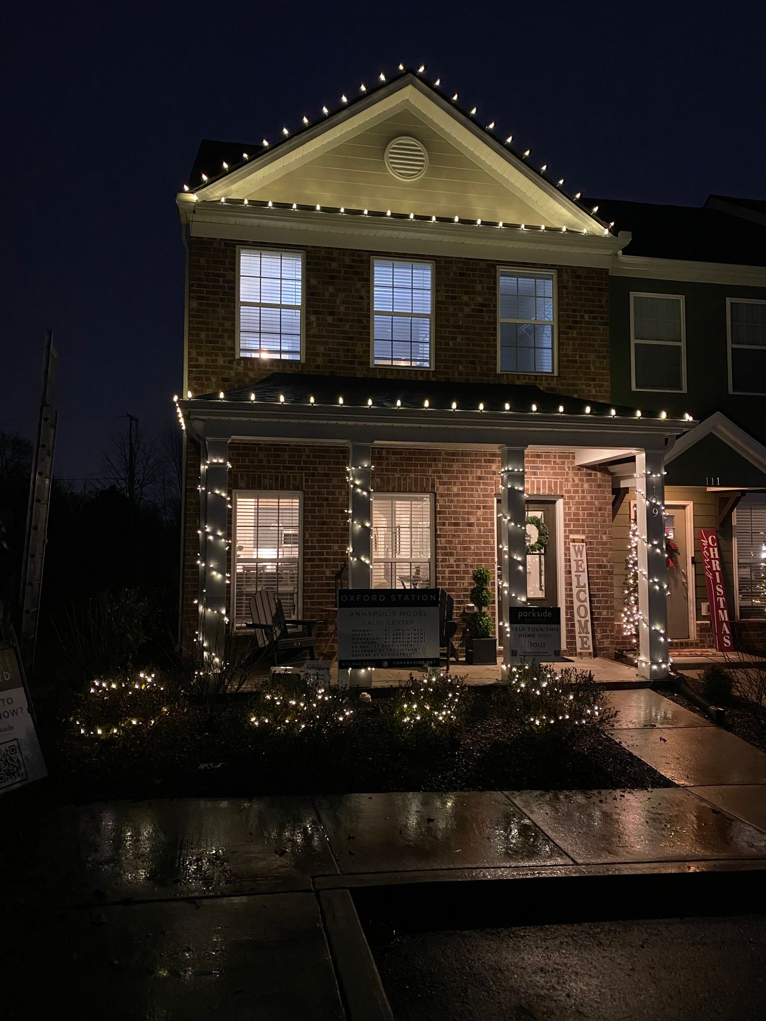 A two-story brick house at night decorated with white Christmas lights. Lights outline the roof, porch, and bushes in front.