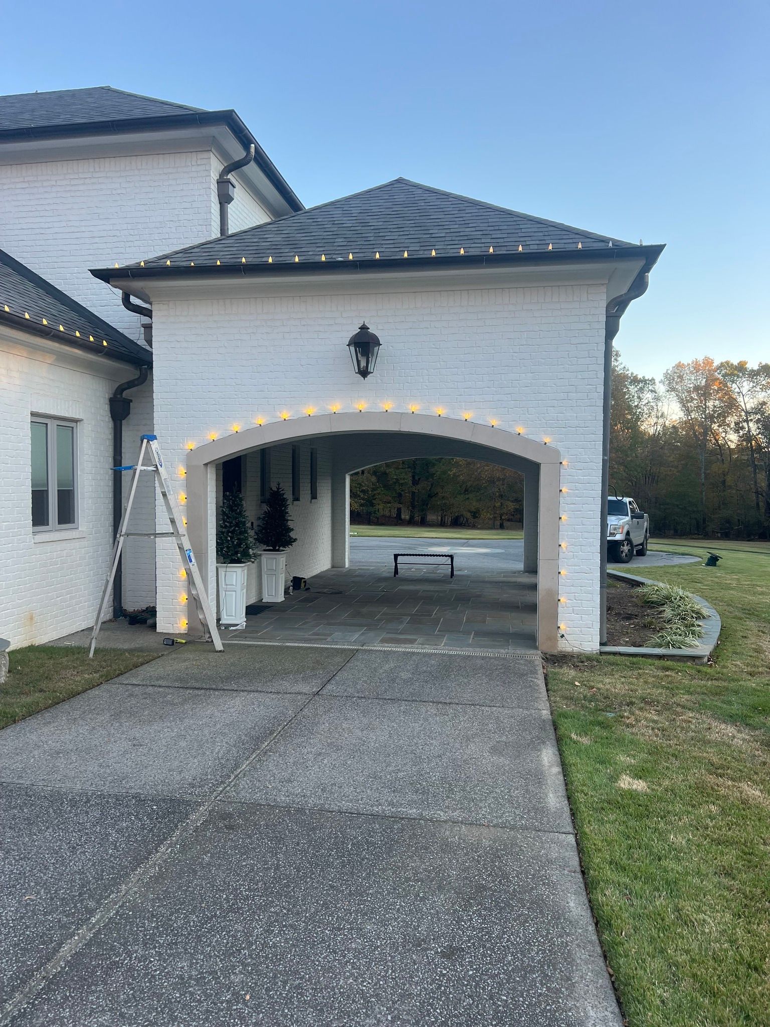 A brick house with Christmas lights lining the roof and arched entrance. A ladder and decorations are present.