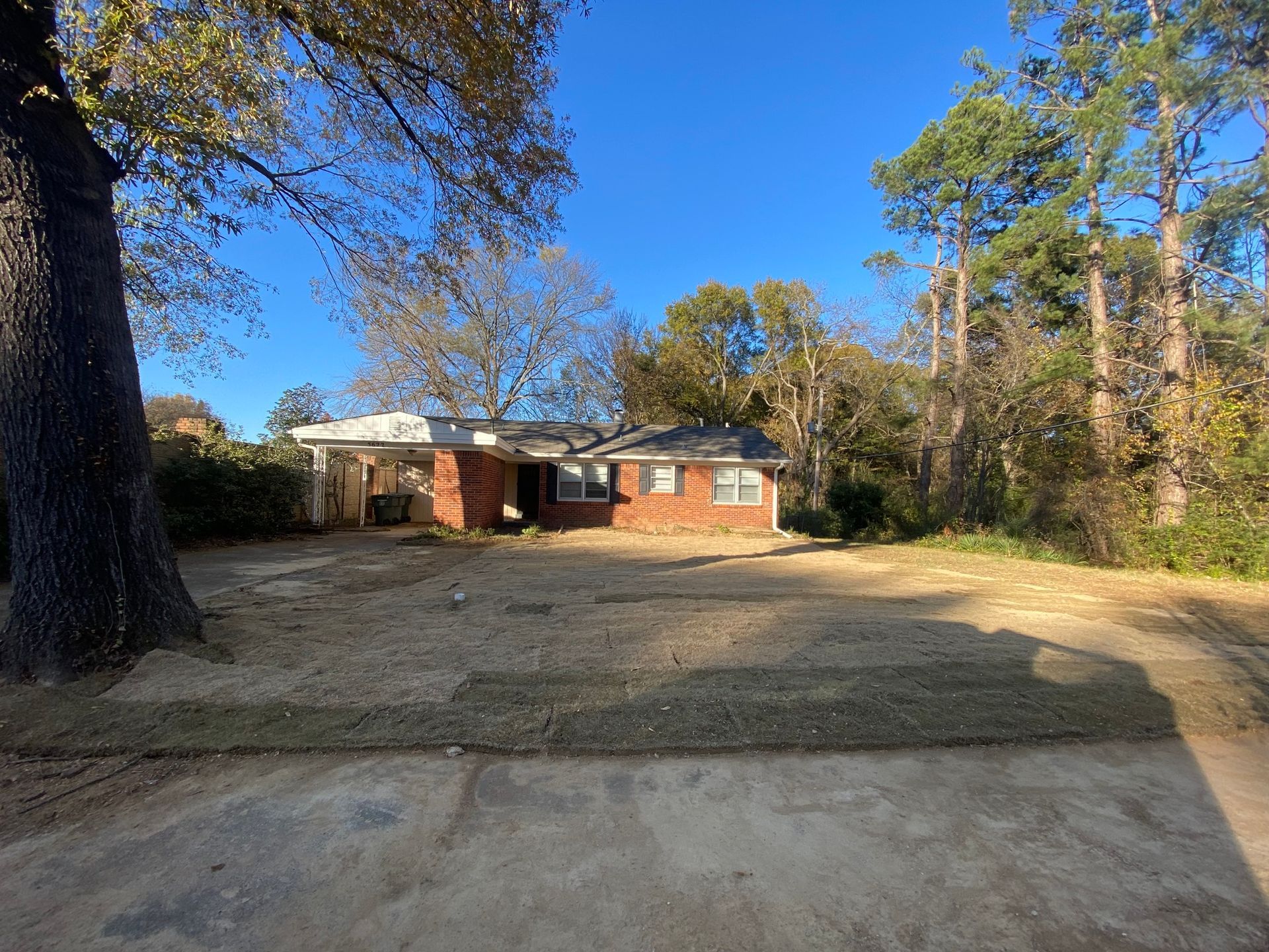 A single-story brick house with a carport, partially surrounded by trees and a newly sodded lawn under a blue sky.
