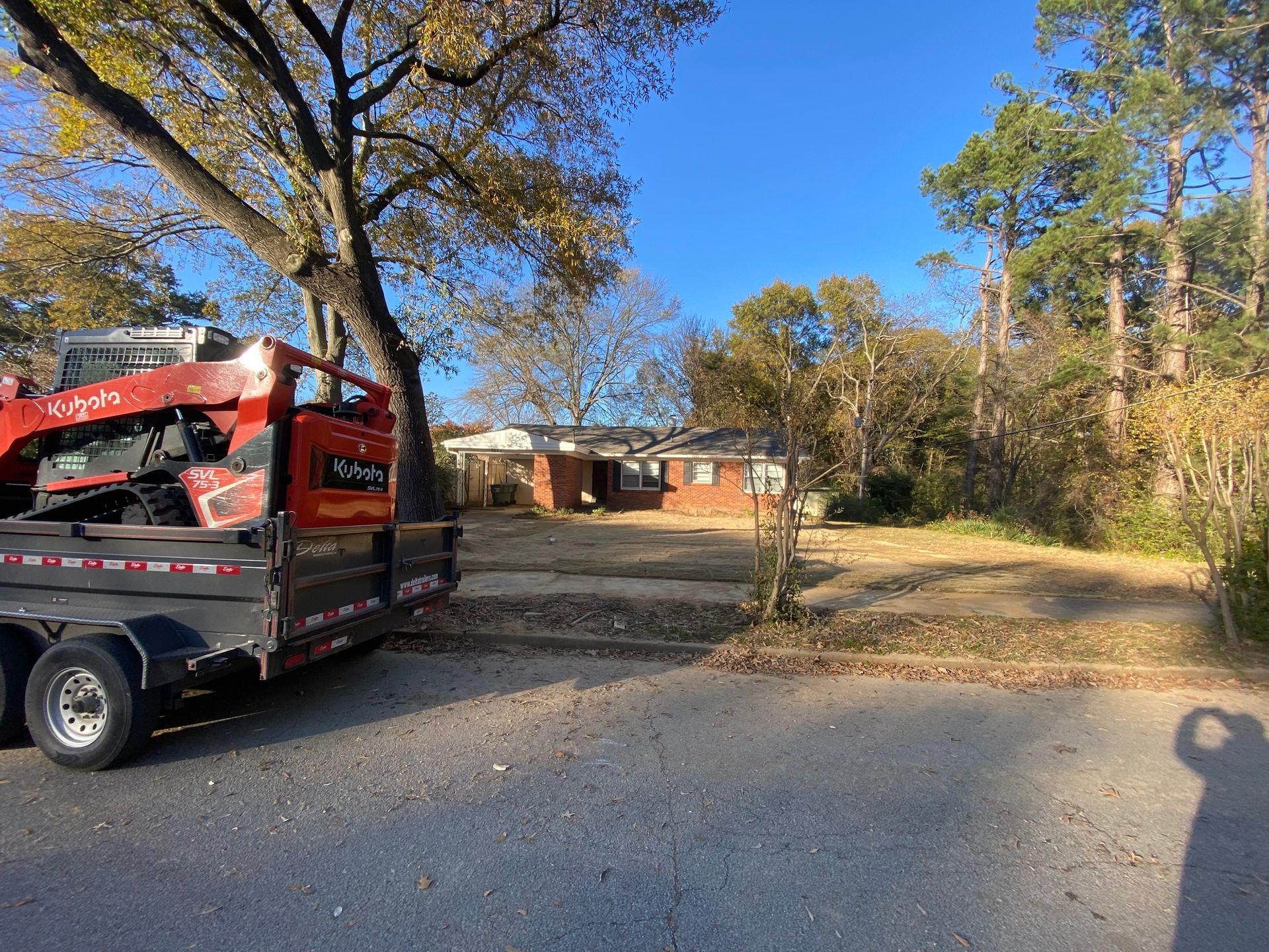 A red trailer with equipment parked in front of a single-story brick house on a sunny day. Bare trees and foliage surround the house.