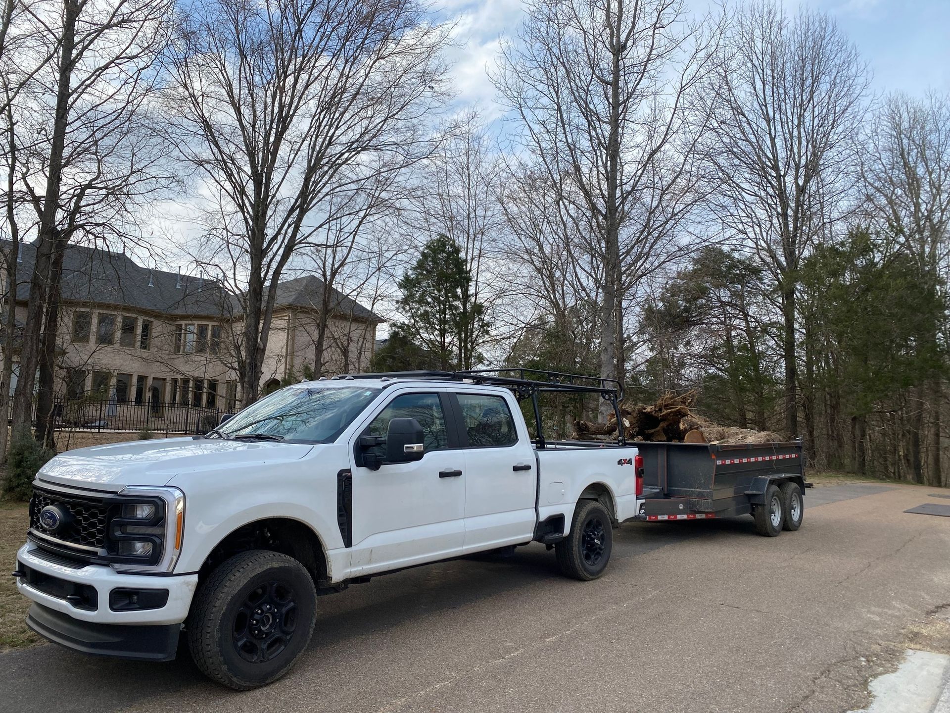 White pickup truck towing a trailer loaded with wood, parked on a road in front of a large house.