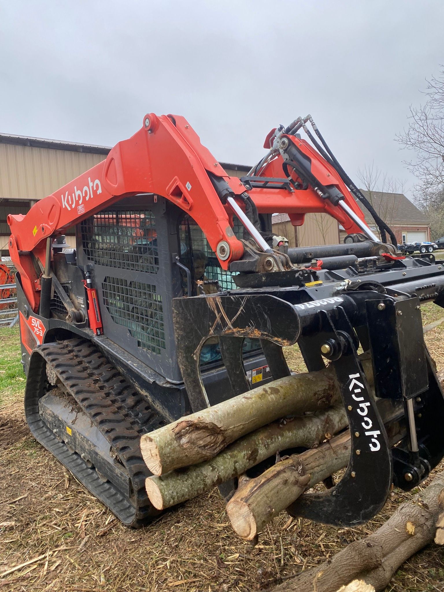 A Kubota track skid steer, orange and black, gripping logs. It is outdoors on a cloudy day.