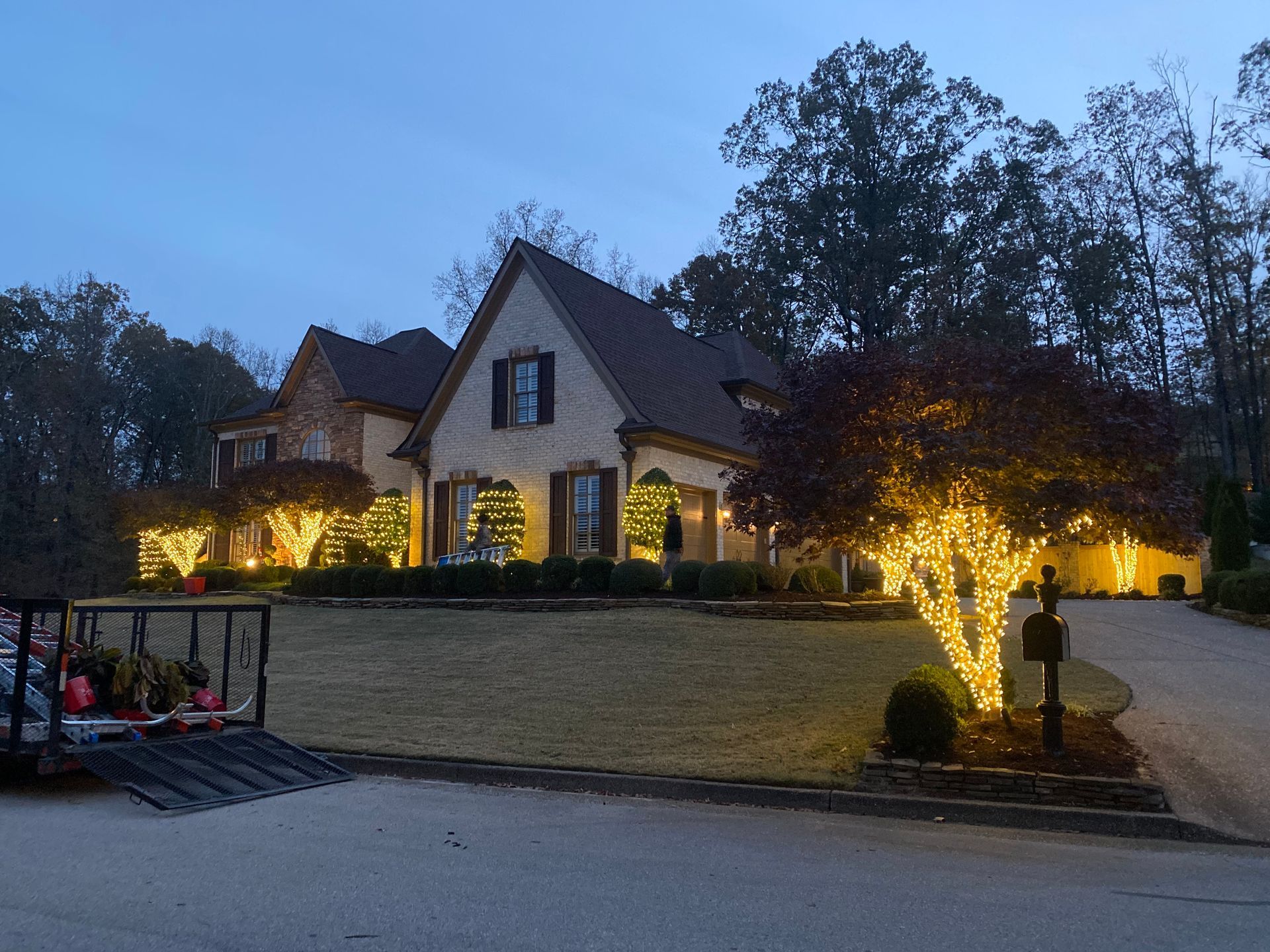 A house with Christmas lights decorating the trees and bushes in the front yard at dusk. A trailer is in the foreground.