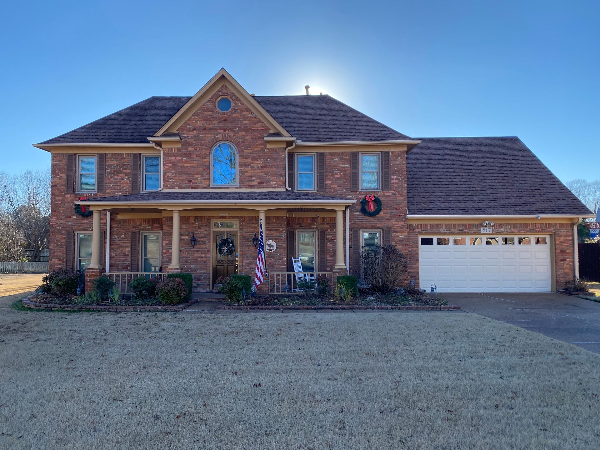 Two-story brick house with a covered porch, garage, and brown roof. The yard is brown grass, and the sky is clear.